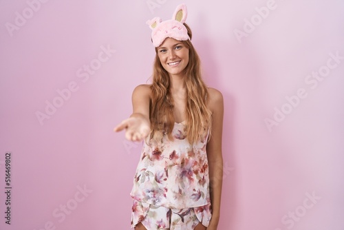Young caucasian woman wearing sleep mask and pajama smiling cheerful offering palm hand giving assistance and acceptance.