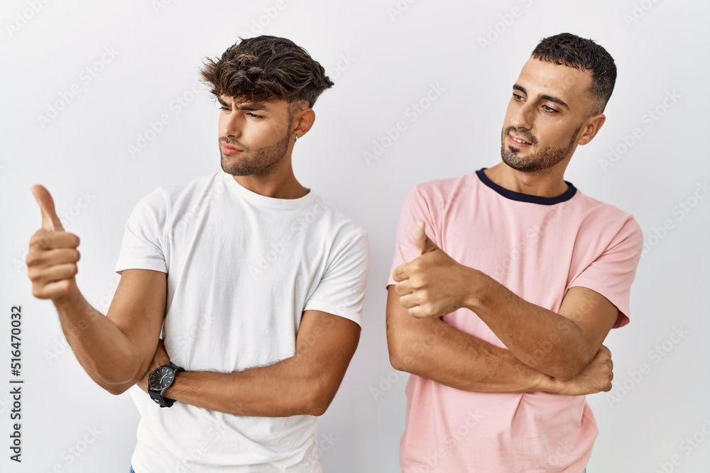 Young gay couple standing over isolated background looking proud, smiling doing thumbs up gesture to the side Young gay couple standing over isolated background looking proud, smiling doing thumbs up gesture to the side