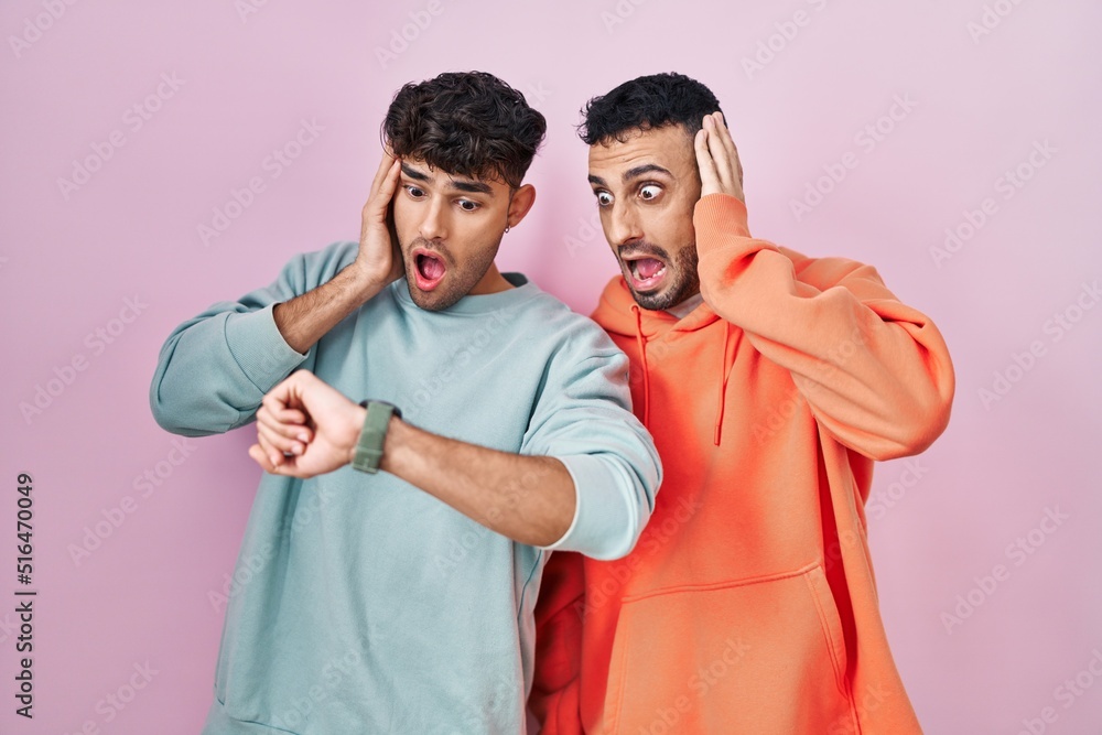 Young hispanic gay couple standing over pink background looking at the watch time worried, afraid of getting late Young hispanic gay couple standing over pink background looking at the watch time worried, afraid of getting late