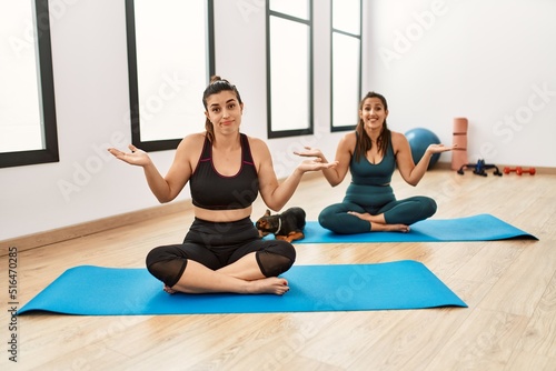 Two hispanic women friends training at the gym sitting on yoga mat with dog clueless and confused expression with arms and hands raised. doubt concept.