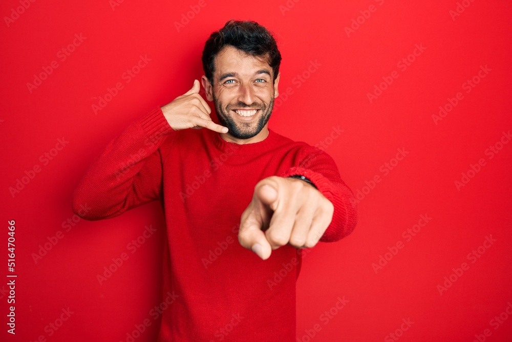 Handsome man with beard wearing casual red sweater smiling doing talking on the telephone gesture and pointing to you. call me. Handsome man with beard wearing casual red sweater smiling doing talking on the telephone gesture and pointing to you. call me.