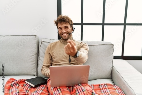 Young handsome man with beard wearing operator headset working from home beckoning come here gesture with hand inviting welcoming happy and smiling