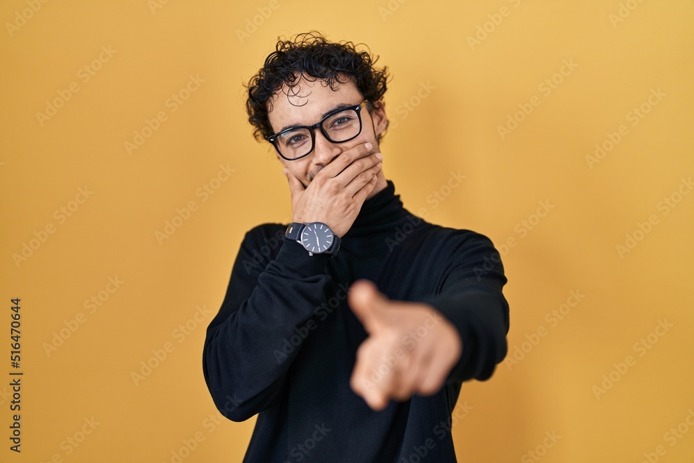 Hispanic man standing over yellow background laughing at you, pointing finger to the camera with hand over mouth, shame expression Hispanic man standing over yellow background laughing at you, pointing finger to the camera with hand over mouth, shame expression