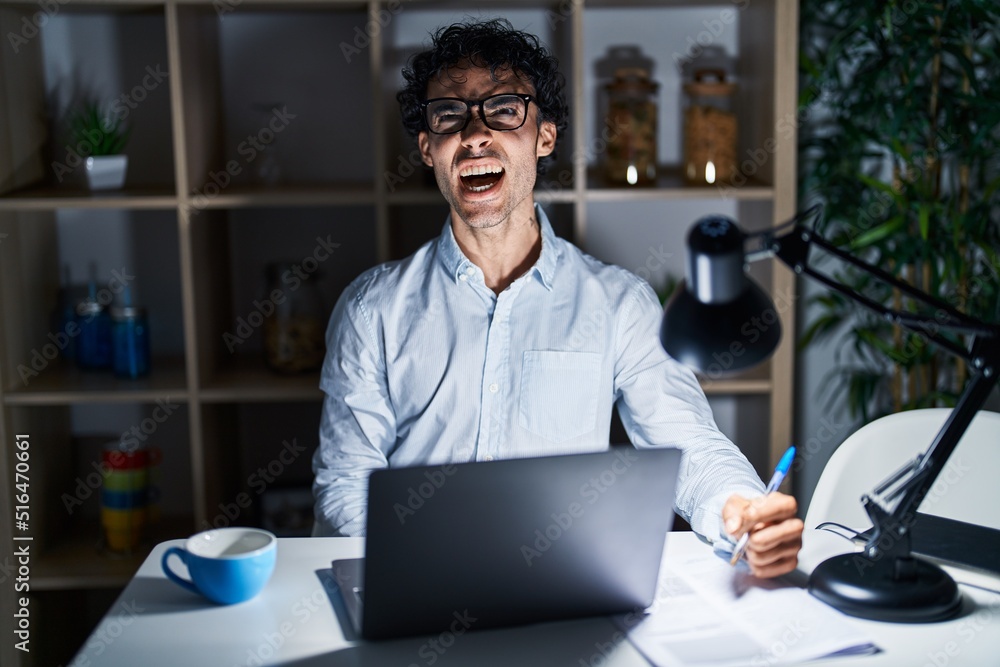 Hispanic man working at the office at night angry and mad screaming frustrated and furious, shouting with anger. rage and aggressive concept. Hispanic man working at the office at night angry and mad screaming frustrated and furious, shouting with anger. rage and aggressive concept.