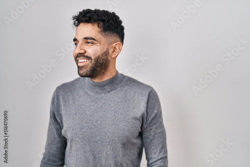 Hispanic man with beard standing over white background looking away to side with smile on face, natural expression. laughing confident.