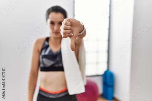 Young brunette woman wearing sportswear and towel at the gym looking unhappy and angry showing rejection and negative with thumbs down gesture. bad expression.