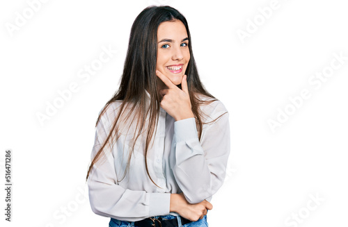 Young brunette teenager wearing business white shirt looking confident at the camera smiling with crossed arms and hand raised on chin. thinking positive.