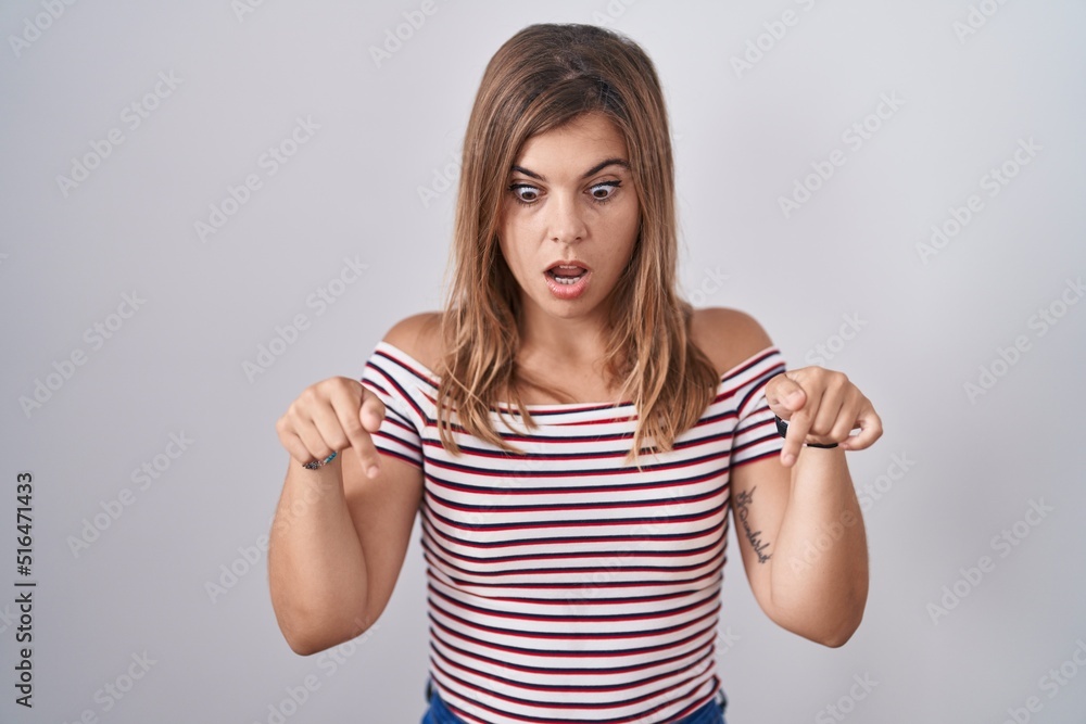 Young hispanic woman standing over isolated background pointing down with fingers showing advertisement, surprised face and open mouth Young hispanic woman standing over isolated background pointing down with fingers showing advertisement, surprised face and open mouth