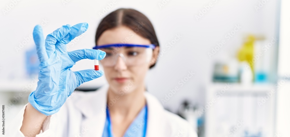Young hispanic woman wearing scientist uniform holding pill at laboratory Young hispanic woman wearing scientist uniform holding pill at laboratory