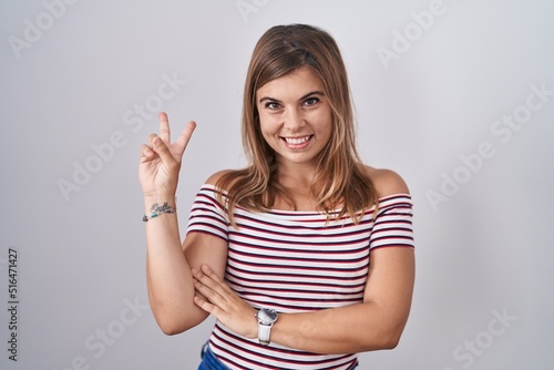 Young hispanic woman standing over isolated background smiling with happy face winking at the camera doing victory sign with fingers. number two.