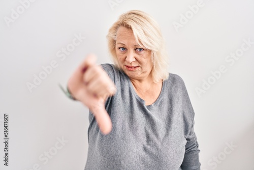 Middle age caucasian woman standing over white background looking unhappy and angry showing rejection and negative with thumbs down gesture. bad expression.