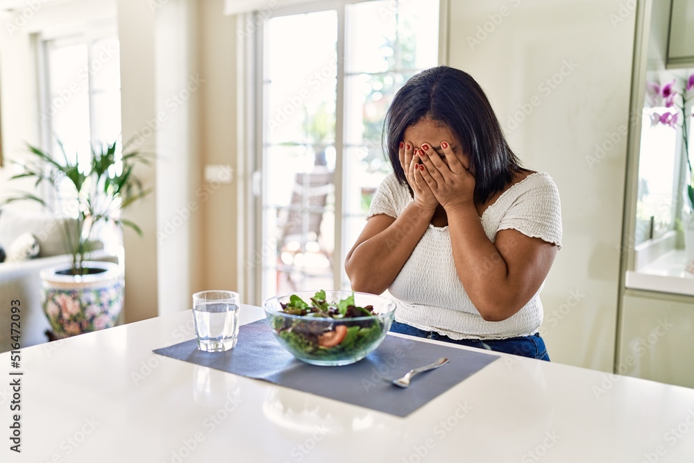 Young hispanic woman eating healthy salad at home with sad expression covering face with hands while crying. depression concept.