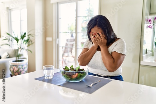 Young hispanic woman eating healthy salad at home with sad expression covering face with hands while crying. depression concept.