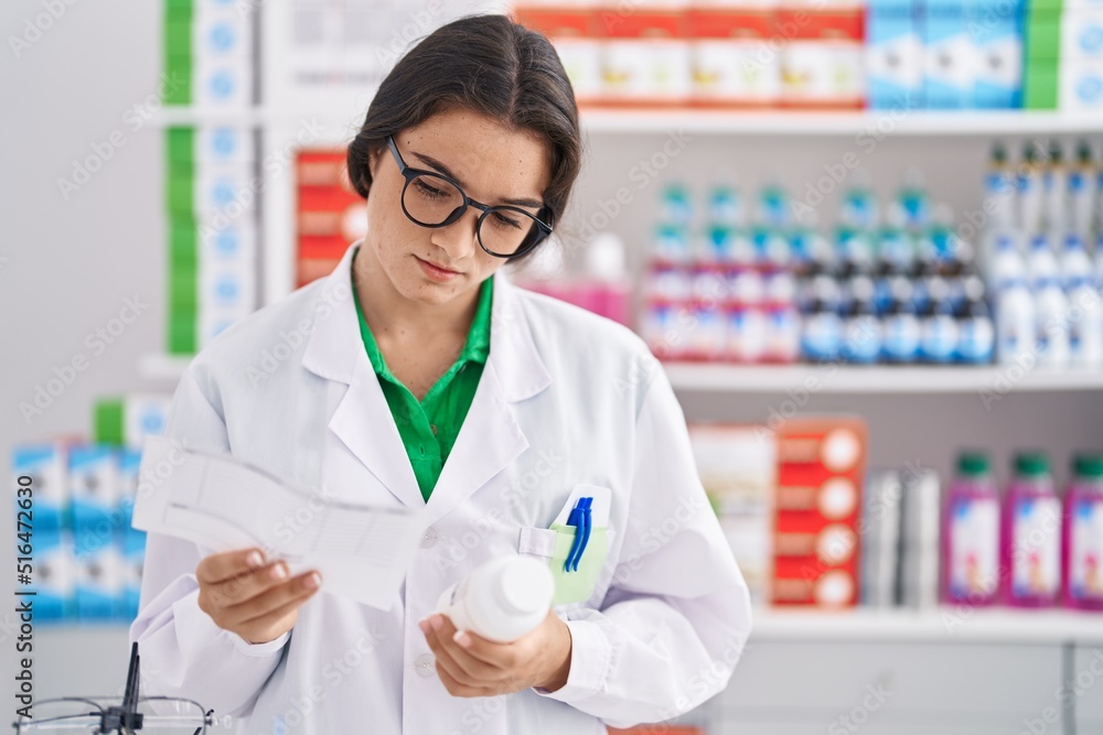 Young hispanic woman pharmacist holding prescription and pills bottle at pharmacy