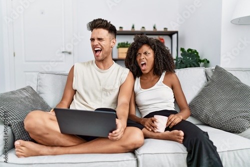 Young interracial couple using laptop at home sitting on the sofa angry and mad screaming frustrated and furious, shouting with anger. rage and aggressive concept.