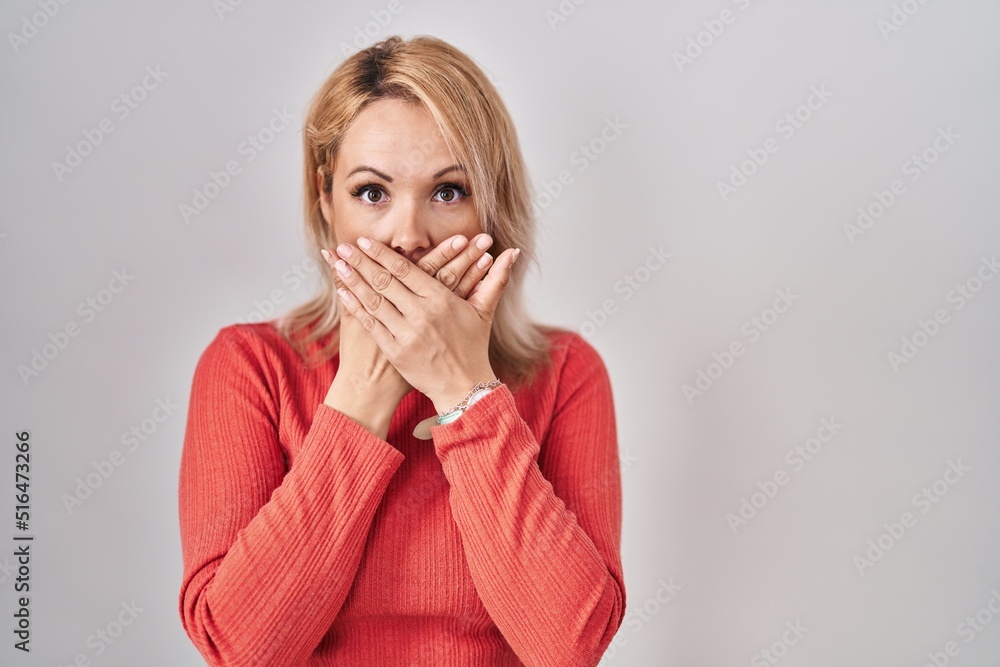 Blonde woman standing over isolated background shocked covering mouth with hands for mistake. secret concept. Blonde woman standing over isolated background shocked covering mouth with hands for mistake. secret concept.