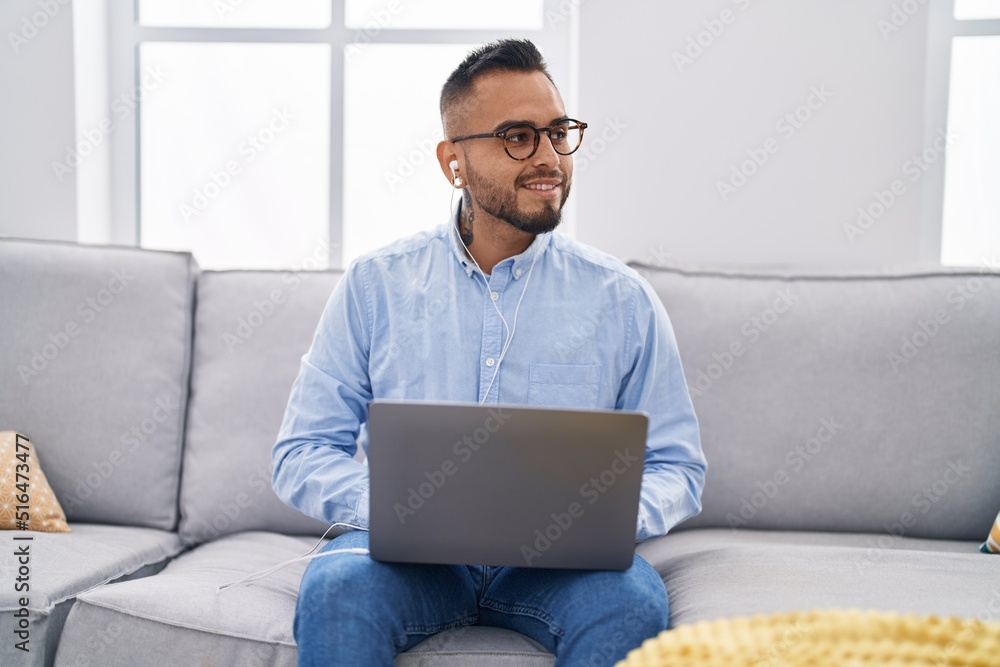 Young hispanic man using laptop and earphones sitting on sofa at home
