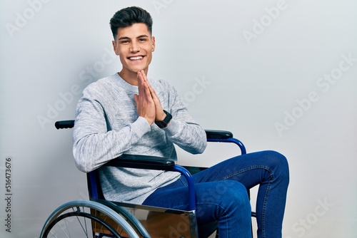 Young hispanic man sitting on wheelchair praying with hands together asking for forgiveness smiling confident.