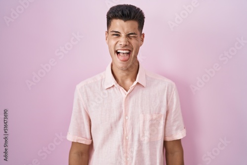 Young hispanic man standing over pink background sticking tongue out happy with funny expression. emotion concept.