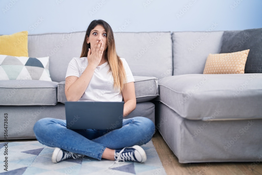 Young brunette woman sitting on the floor at home using laptop covering mouth with hand, shocked and afraid for mistake. surprised expression Young brunette woman sitting on the floor at home using laptop covering mouth with hand, shocked and afraid for mistake. surprised expression