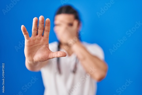 Hispanic man with long hair standing over blue background covering eyes with hands and doing stop gesture with sad and fear expression. embarrassed and negative concept.