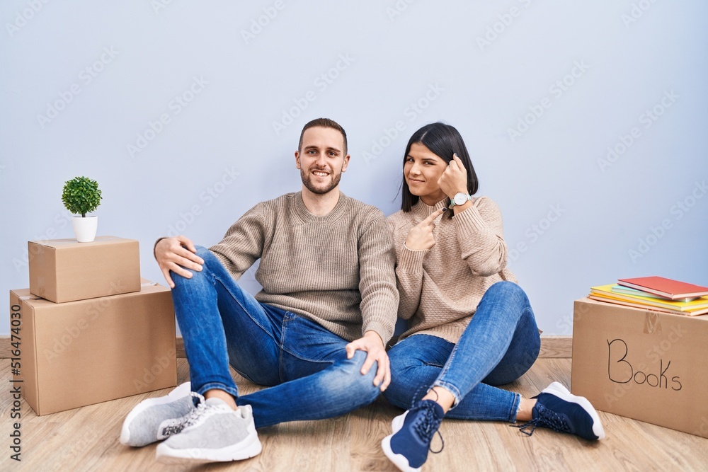 Young couple moving to a new home in hurry pointing to watch time, impatience, looking at the camera with relaxed expression Young couple moving to a new home in hurry pointing to watch time, impatience, looking at the camera with relaxed expression