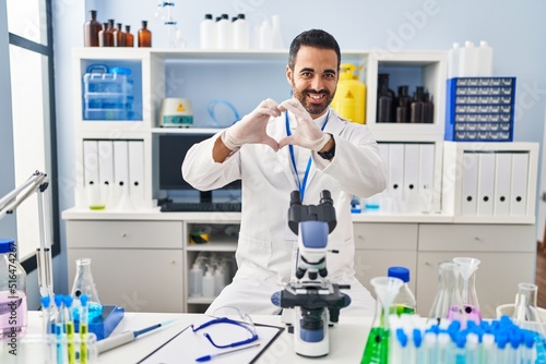 Young hispanic man with beard working at scientist laboratory smiling in love doing heart symbol shape with hands. romantic concept.