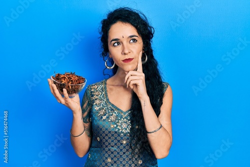 Young woman wearing bindi and traditional kurta dress holding bowl of star anise serious face thinking about question with hand on chin, thoughtful about confusing idea photo