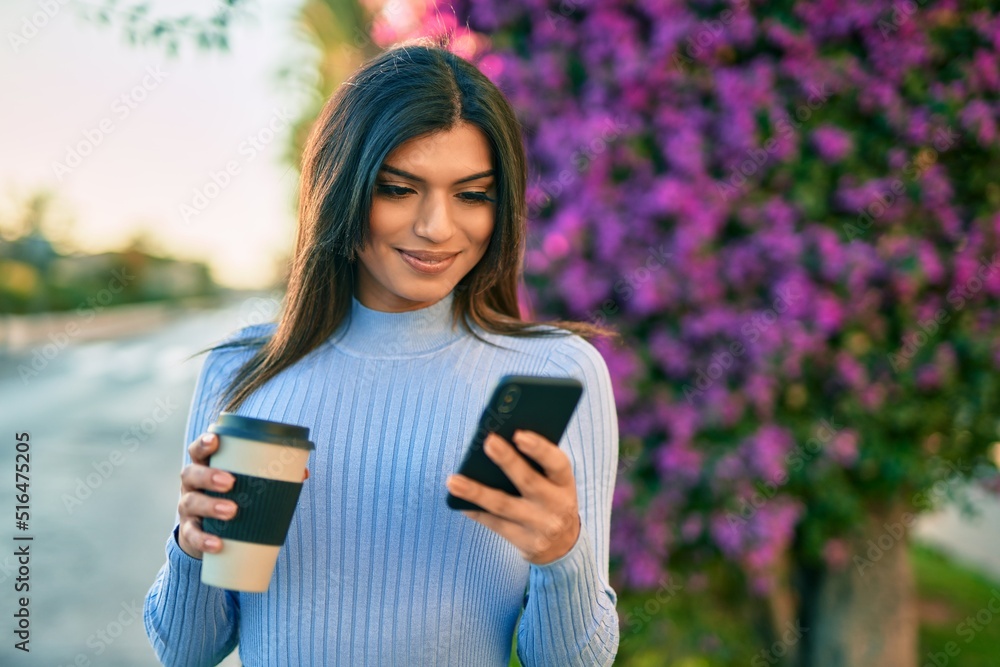 Young hispanic woman using smartphone and drinking a cup of coffee by the park