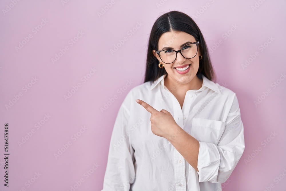 Young brunette woman standing over pink background cheerful with a smile of face pointing with hand and finger up to the side with happy and natural expression on face Young brunette woman standing over pink background cheerful with a smile of face pointing with hand and finger up to the side with happy and natural expression on face
