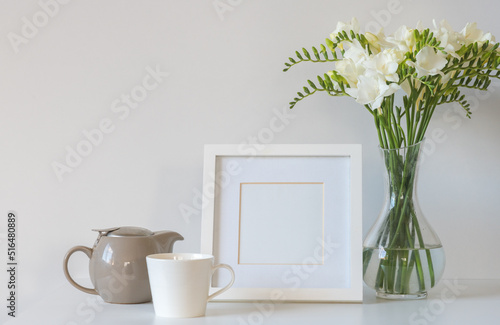 White freesia flowers in glass vase with blank square picture frame, tea cup and teapot against white background (selective focus) © Natalie Board