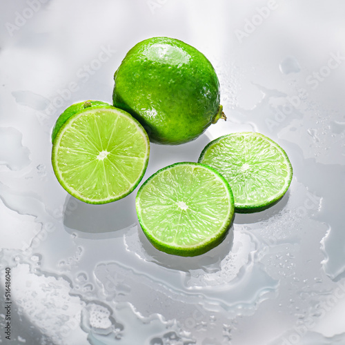The green lime is blank with the cut lime slice showing the inside of the wet lemon pulp on a clear glass surface, reflecting the shadows of the lime and the wet water, giving it its freshness.