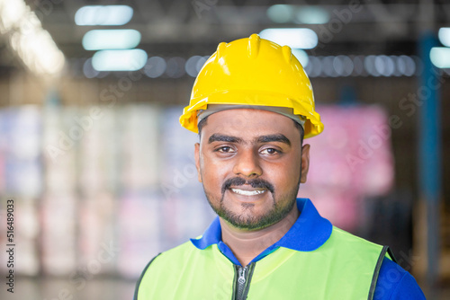 Portrait of Worker wearing hardhat and safety vest standing in the warehouse, Cheerful man looking at camera with blurred background and copy space © JU.STOCKER