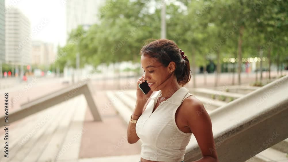 Lovely tanned woman with ponytail wearing white top is using mobile phone. Girl talking on smartphone going down the stairs on the cityscape background Lovely tanned woman with ponytail wearing white top is using mobile phone. Girl talking on smartphone going down the stairs on the cityscape background