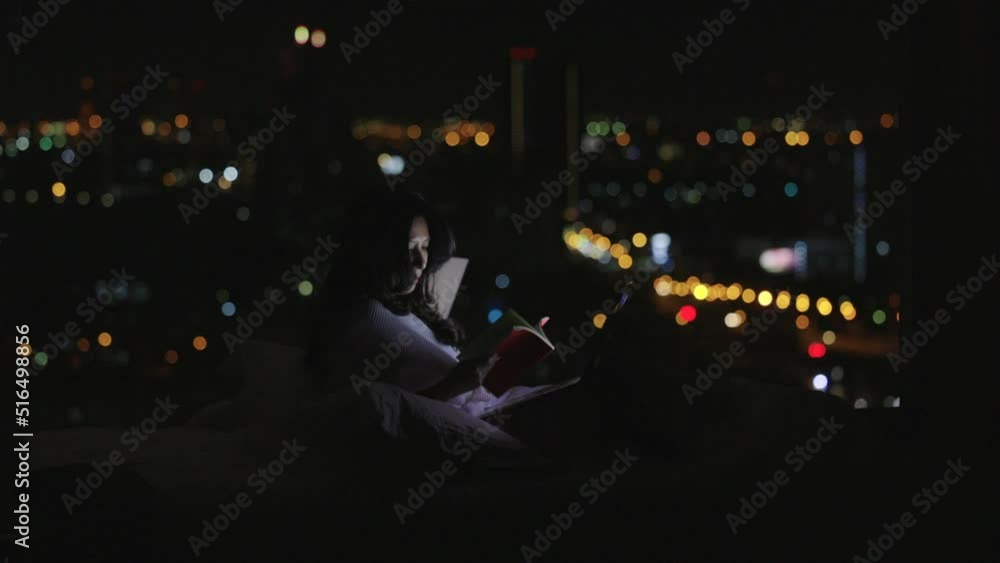 Beautiful Asian woman in bathrobe sitting in white cozy bed with open book and using laptop with bright light screen in the late night at home with panorama window and Blurred bokeh, night city view. Beautiful Asian woman in bathrobe sitting in white cozy bed with open book and using laptop with bright light screen in the late night at home with panorama window and Blurred bokeh, night city view.