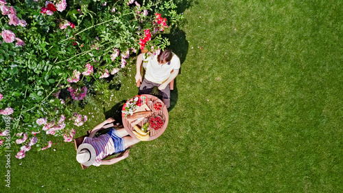 Young couple enjoying food and drinks in beautiful roses garden on romantic date, aerial top view from above of man and woman eating and drinking together outdoors in park 