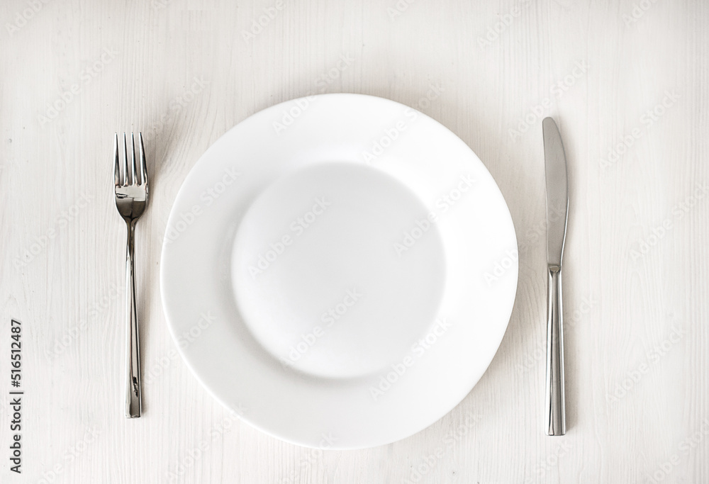 White clean plate and stainless knife and fork on light wooden table. Served cutlery. Top view White clean plate and stainless knife and fork on light wooden table. Served cutlery. Top view