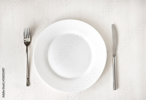 White clean plate and stainless knife and fork on light wooden table. Served cutlery. Top view