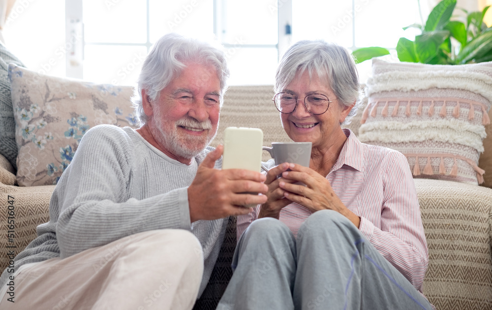 Smiling serene beautiful caucasian senior couple sitting on the floor at home using smart phone, old people and new technologies concept Smiling serene beautiful caucasian senior couple sitting on the floor at home using smart phone, old people and new technologies concept