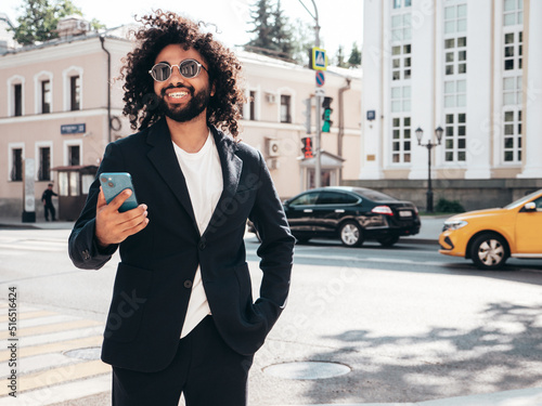 Handsome smiling hipster model.Sexy unshaven Arabian man dressed in summer suit jacket clothes.Fashion male with long curly hairstyle in the street, using smartphone apps, looking at cellphone screen