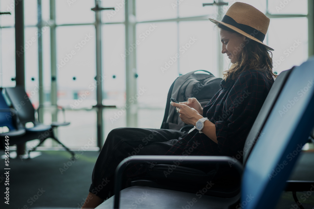 Young beautiful woman traveler in a hat with a backpack using a smartphone while sitting at the airport in the departure area Young beautiful woman traveler in a hat with a backpack using a smartphone while sitting at the airport in the departure area