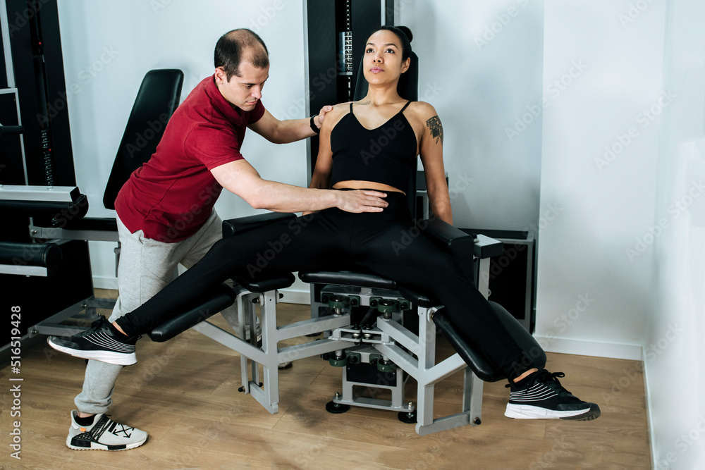 Young, athletic girl using a gym machine with her personal trainer to strengthen her legs and buttocks.
