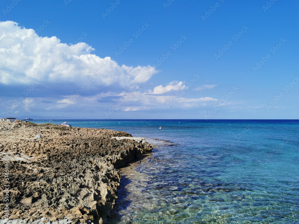 Stone shore of solidified lava, clear water against a blue sky with clouds.
