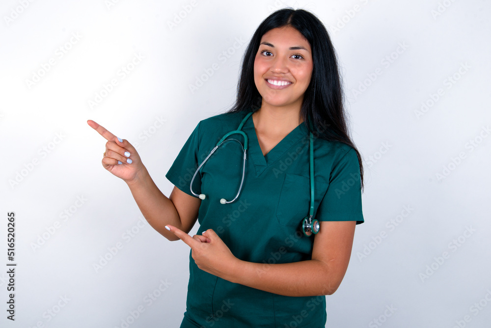 Doctor hispanic woman wearing surgeon uniform over white wall indicating finger empty space showing best low prices, looking at the camera