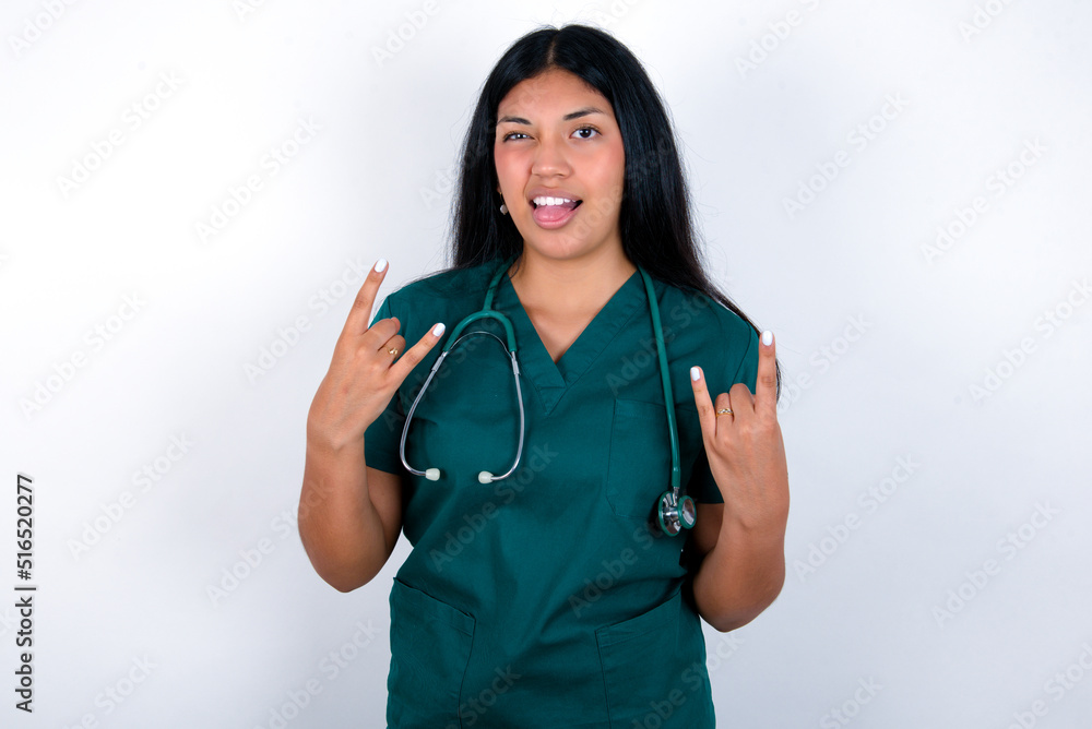 Doctor hispanic woman wearing surgeon uniform over white wall making rock hand gesture and showing tongue Doctor hispanic woman wearing surgeon uniform over white wall making rock hand gesture and showing tongue