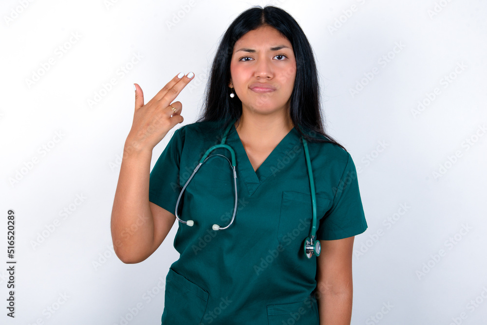 Unhappy Doctor hispanic woman wearing surgeon uniform over white wall makes suicide gesture and imitates gun with hand, curves lips, keeps two fingers on temple Unhappy Doctor hispanic woman wearing surgeon uniform over white wall makes suicide gesture and imitates gun with hand, curves lips, keeps two fingers on temple
