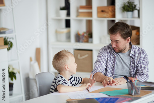 Surprised young father with frowning forehead sitting at table and discussing creative school task with son