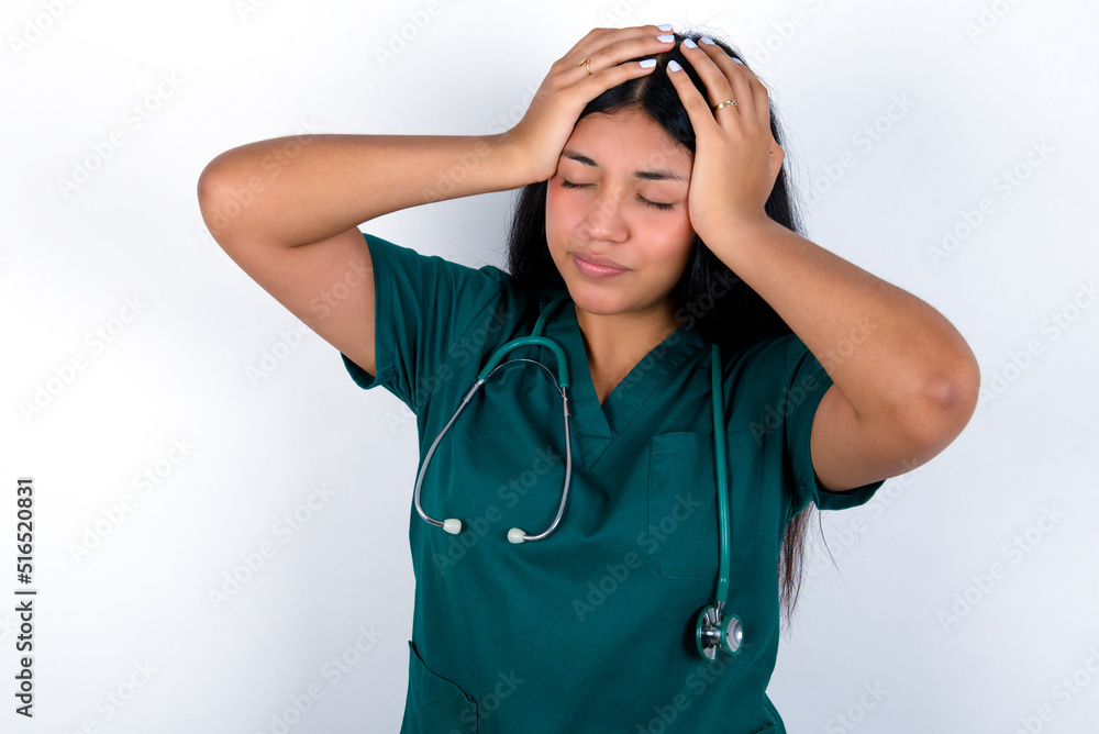 Doctor hispanic woman wearing surgeon uniform over white wall suffering from strong headache desperate and stressed because of overwork. Depression and pain concept. Doctor hispanic woman wearing surgeon uniform over white wall suffering from strong headache desperate and stressed because of overwork. Depression and pain concept.