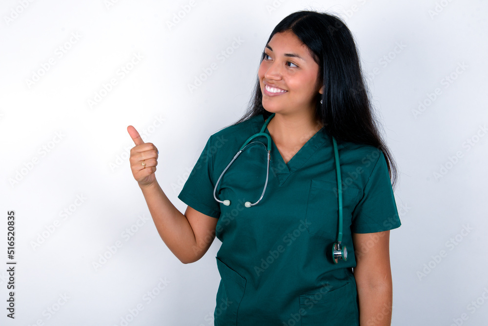 Doctor hispanic woman wearing surgeon uniform over white wall Looking proud, smiling doing thumbs up gesture to the side. Good job! Doctor hispanic woman wearing surgeon uniform over white wall Looking proud, smiling doing thumbs up gesture to the side. Good job!