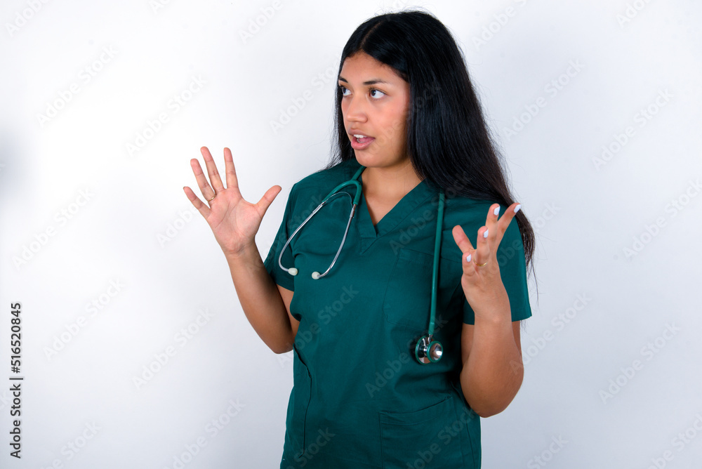 Doctor hispanic woman wearing surgeon uniform over white wall shouts loud, keeps eyes opened and hands tense. Doctor hispanic woman wearing surgeon uniform over white wall shouts loud, keeps eyes opened and hands tense.